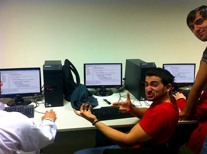 A university computer lab: Nikos turns to the camera in front of a row of monitors showing code.